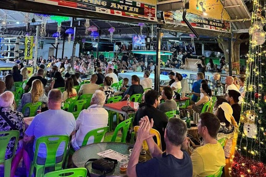 A crowded indoor event with people seated at tables, a stage in the background, and festive decorations. 4