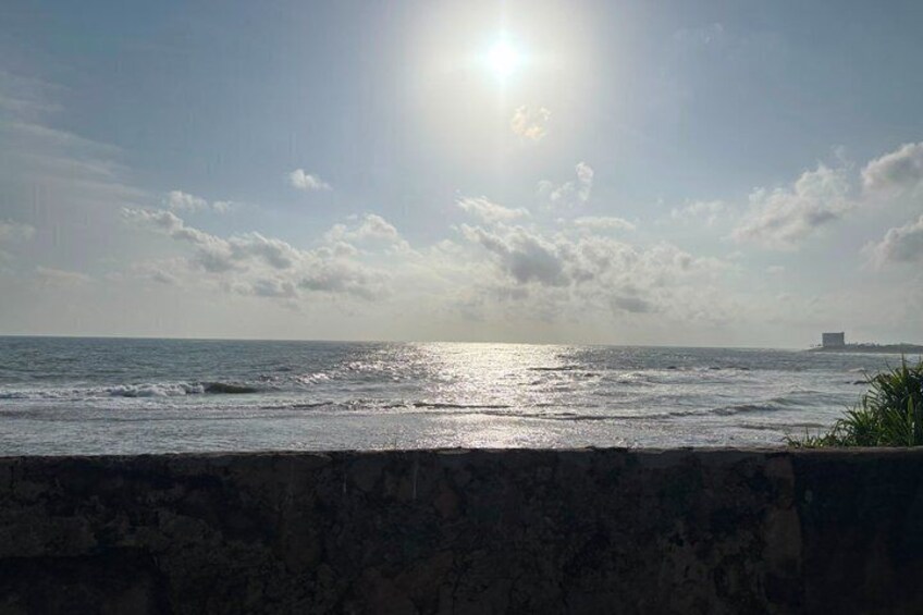 Eine Küstenlandschaft mit Sonnenschein über dem Wasser, eine Steinmauer im Vordergrund und ein Gebäude in der Ferne am Horizont. 4