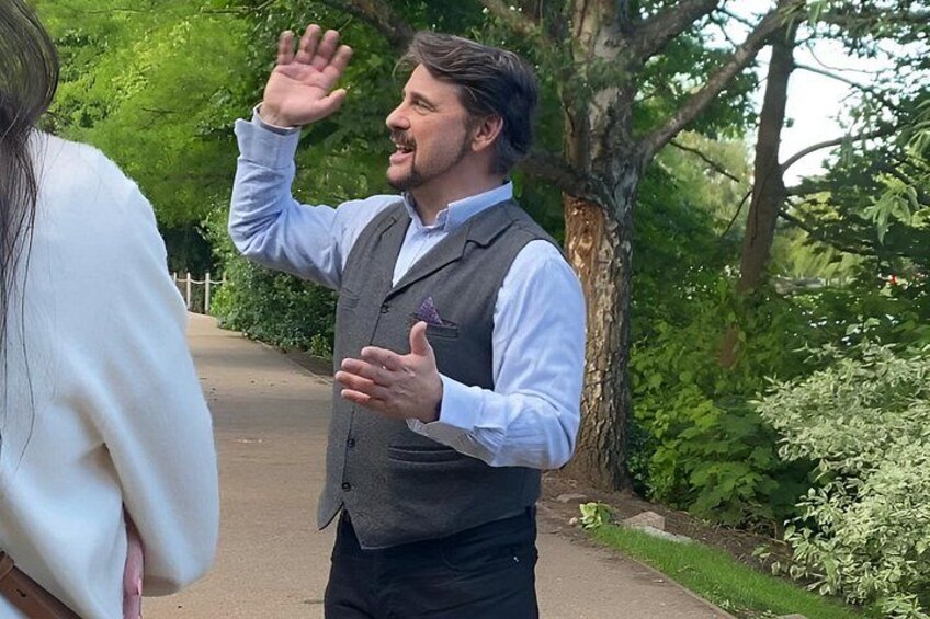 A person in a vest and shirt gesturing, standing on a path with greenery and trees in the background. 2