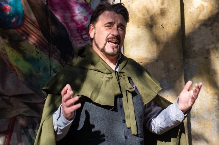 Una persona con un mantello verde e un gilet grigio, che gesticisce con le mani. 1