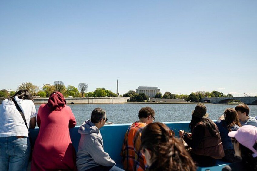 Un groupe de personnes se trouve sur un bateau, près d’un grand bâtiment orné d’une colonne, qui semble être un bâtiment gouvernemental, et ce bâtiment est situé au bord d’une étendue d’eau. 3