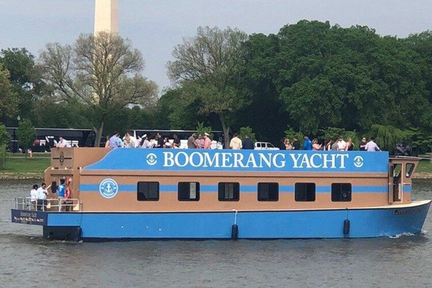 A blue and brown boat named 'BOOMERANG YACHT' on a river with people on board and a large monument in the background. 1