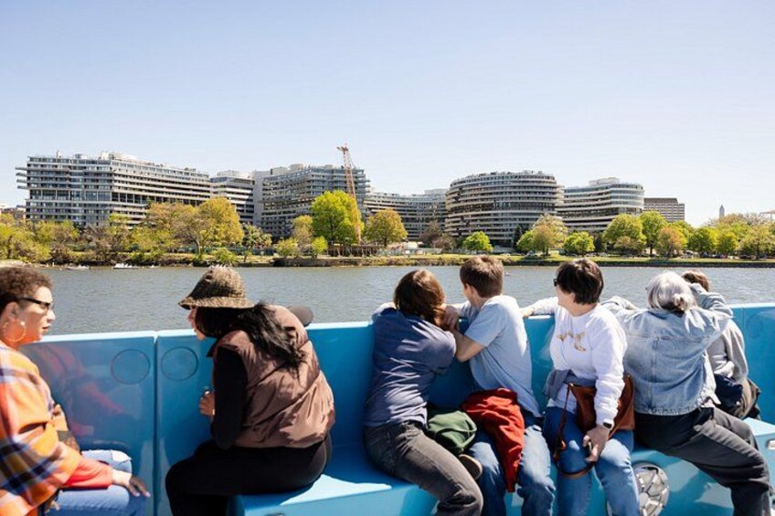 A group of people sitting on a blue bench by a river with buildings in the background. 2