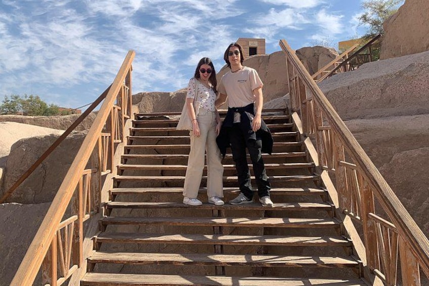 Two people standing on wooden stairs with a metal railing, surrounded by large rock formations. 2