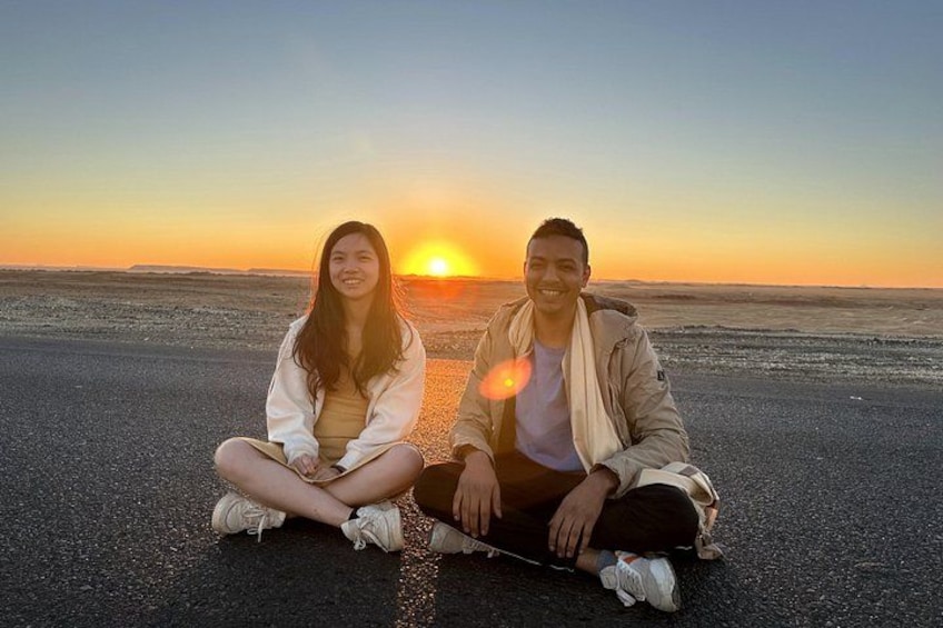Two people sitting on a beach with the sun setting in the background. 3