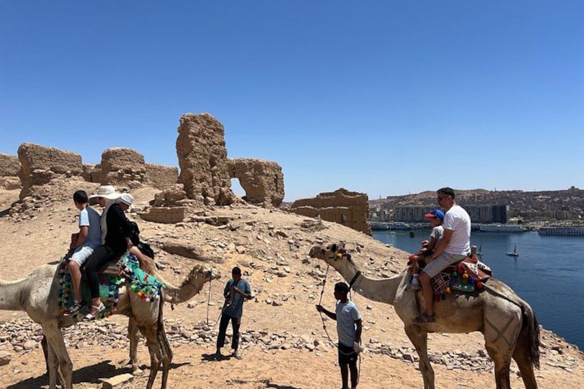 People riding camels on a rocky terrain near a body of water with ancient ruins in the background. 5