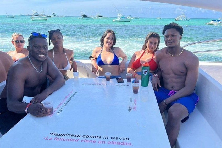 Eine Gruppe Menschen auf einem Boot, vor ihnen ein Tisch, umgeben von Wasser mit Booten im Hintergrund. 5