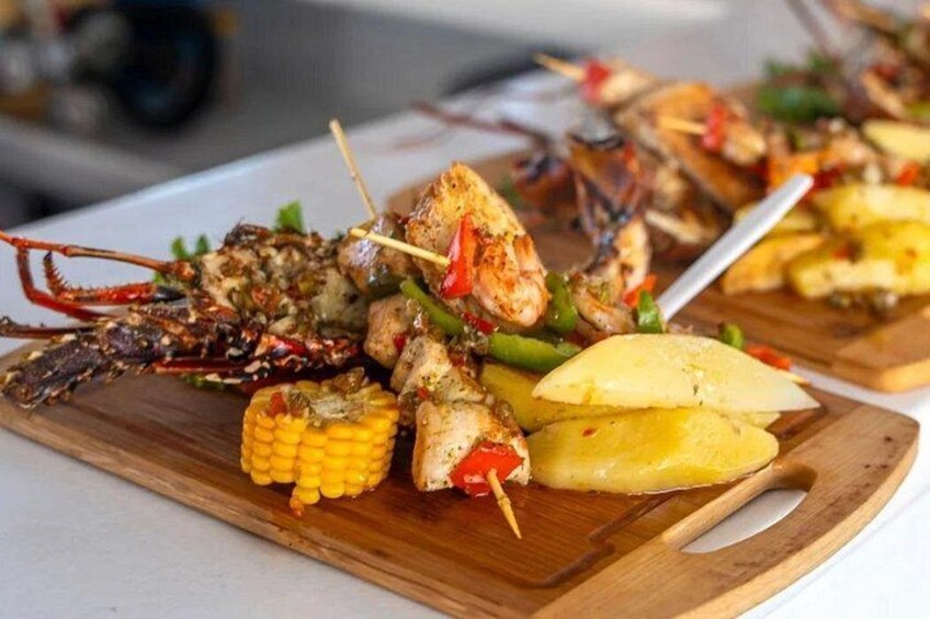 Ein Holzschnittbrett mit gegrilltem Fisch und Meeresfrüchten, darunter Garnelen und Hummer, dazu Maiskolben und Ananasstücke. 3