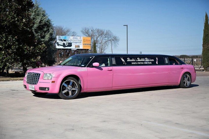 A pink stretch limousine parked in a lot with trees and a billboard in the background. 1