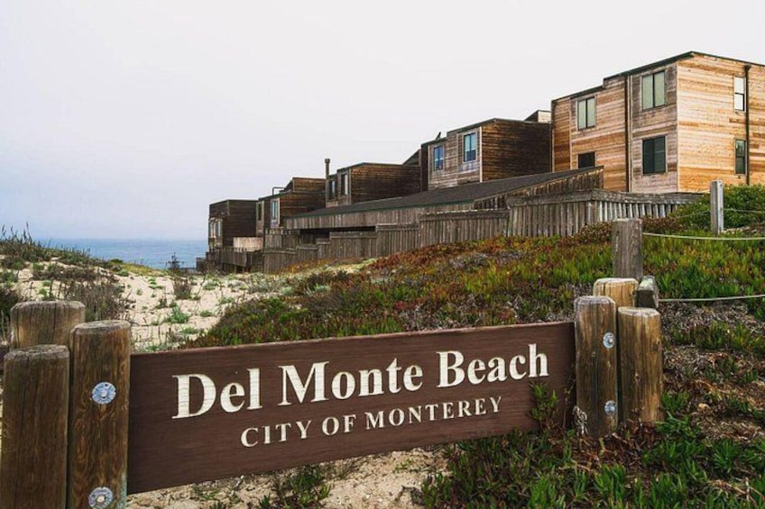 A wooden sign with 'Del Monte Beach City of Monterey' written on it, surrounded by wooden fences and buildings with a beach and ocean in the background. 1