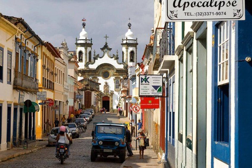 A cobblestone street lined with colorful buildings, a church with white domes, and parked vehicles. 4