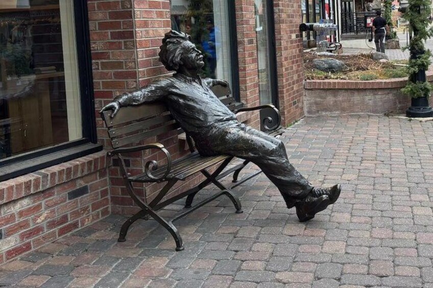 A bronze statue of a man sitting on a bench, a brick building, and a paved walkway. 3
