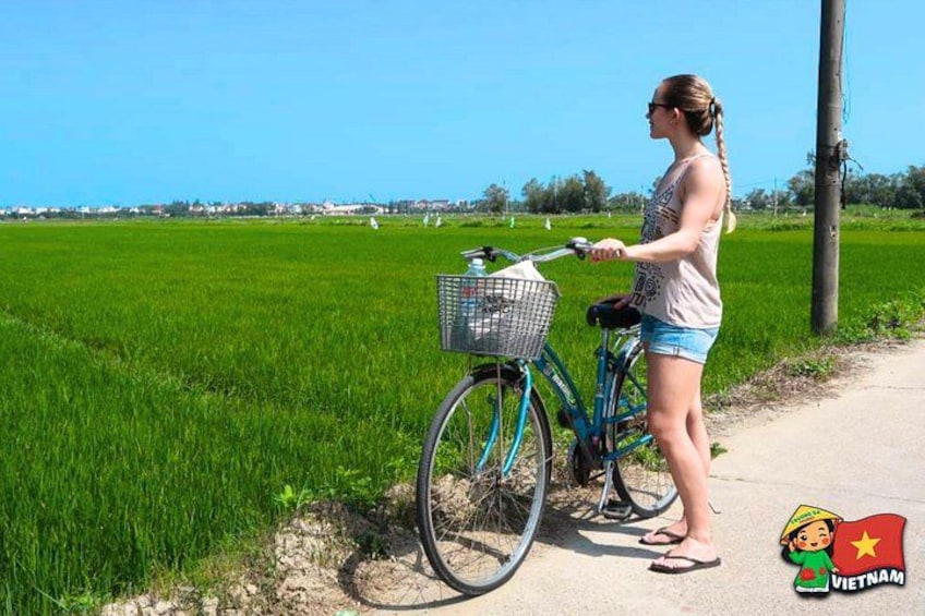 A woman standing next to a blue bicycle with a basket, in a rural setting with green fields and a clear sky. 2