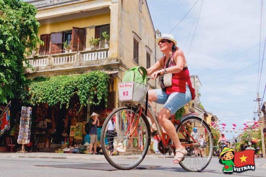 A person riding a bicycle on a street with a building and a shop in the background. 1