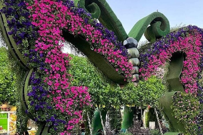 A garden with large sculptures of a butterfly and a caterpillar, surrounded by lush greenery and pink and purple flowers. 1