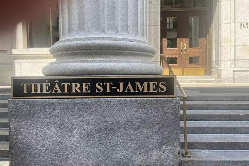 Una placa de piedra con 'THEATRE ST-JAMES' grabado, colocada sobre un pedestal frente a un edificio con una puerta de madera grande. 3
