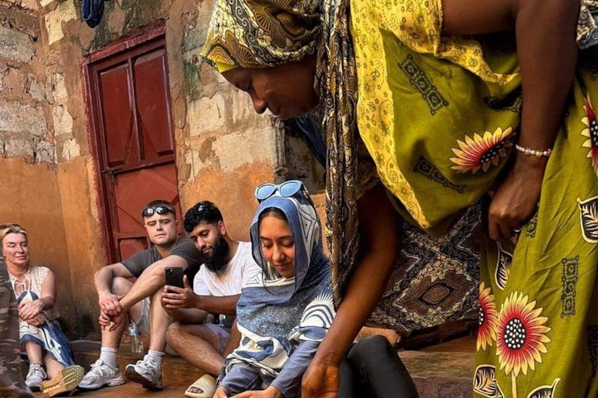 En grupp människor sitter på marken utomhus, med en rustik byggnad i bakgrunden. 3