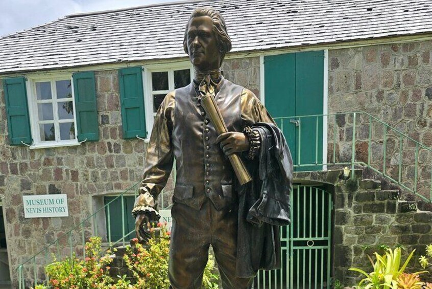 En statue af en historisk person i tidstypisk påklædning, foran en bygning af sten med et skilt, der siger "Museum of Nevis History". 1