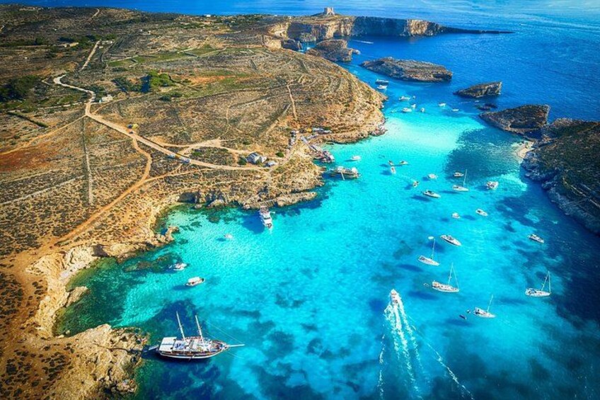 A coastal landscape with clear turquoise waters, boats anchored near the shore, and a lighthouse on a rocky outcrop. 1