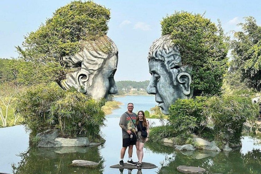 A couple standing between two large stone head sculptures in a body of water surrounded by greenery. 1