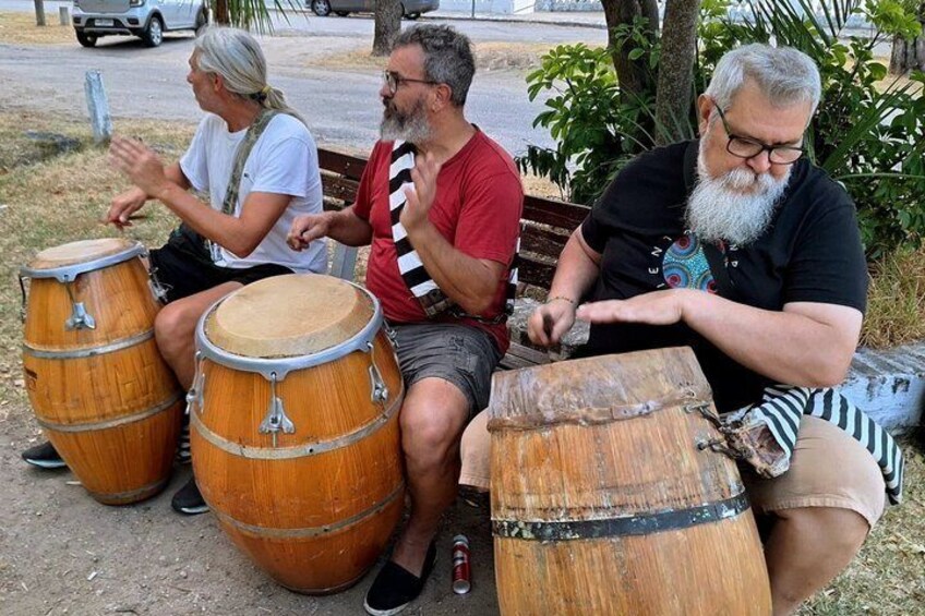 Three people seated on a bench playing large wooden drums. 1