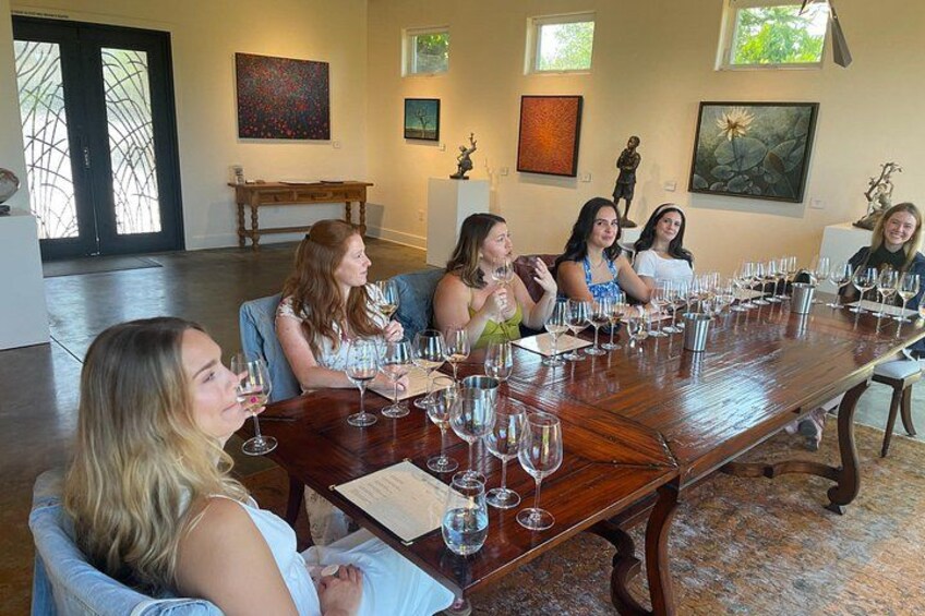 Een groep mensen zit rond een lange houten tafel, met wijnkaraffen. 2
