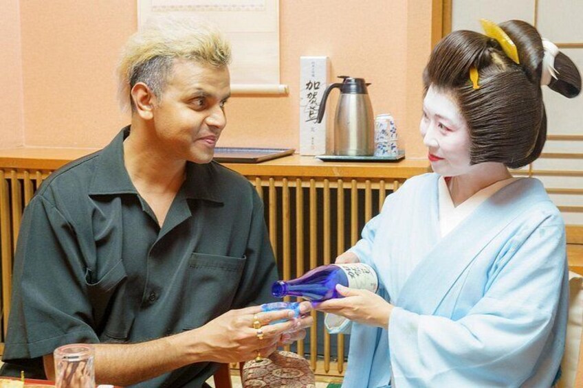 To personer, en i sort skjorte og den anden i traditionelt japansk tøj, sidder ved et bord med et thestel og en kop. 5