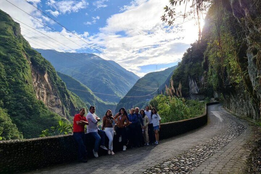 Un groupe de personnes est assis sur un chemin pavé, au bord d’une route de montagne, avec une végétation luxuriante et une vallée en arrière-plan. 4