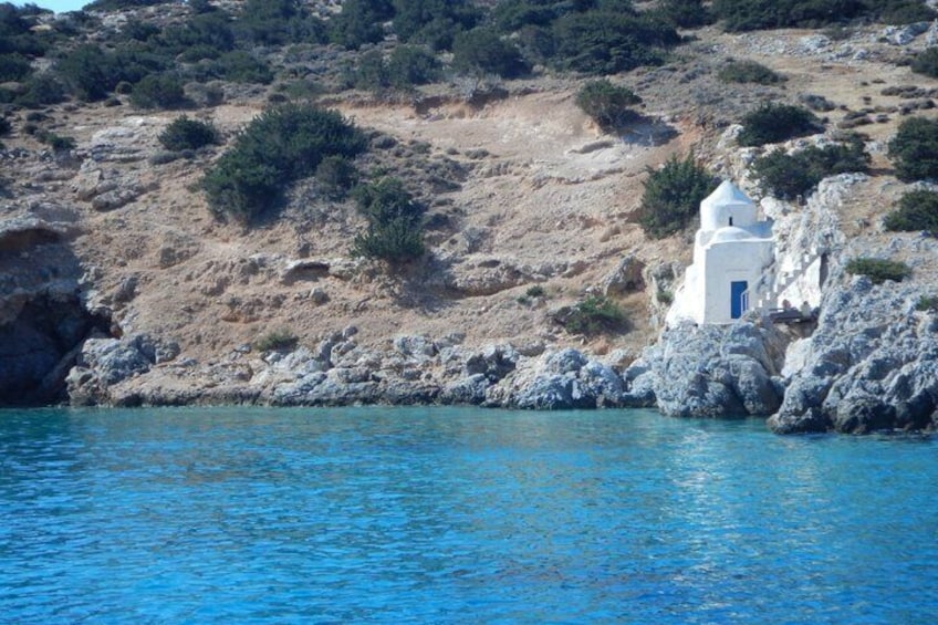 A white church with a blue door on a rocky hillside by a blue sea. 5