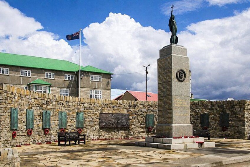 En stenvæg med en bænk, en statue og en bygning med grønt tag. 3