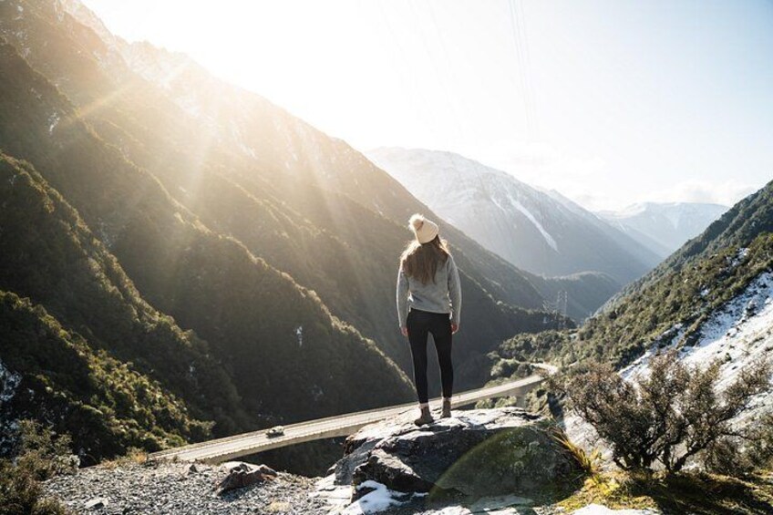 En person står på en klippe med udsigt over et bjergrigt landskab, hvor en vej kan anes i det fjerne. 4