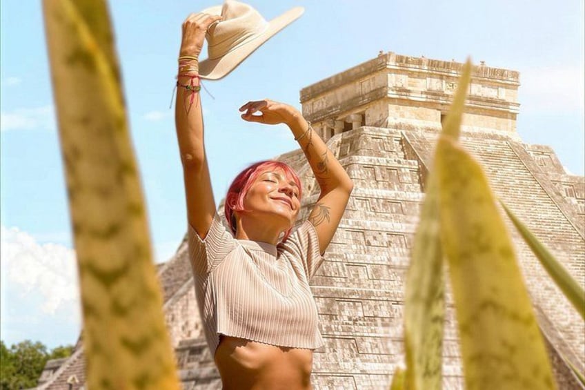 Una persona de cabello rosa, con una camiseta a rayas, sostiene un sombrero frente a una gran pirámide antigua. 1