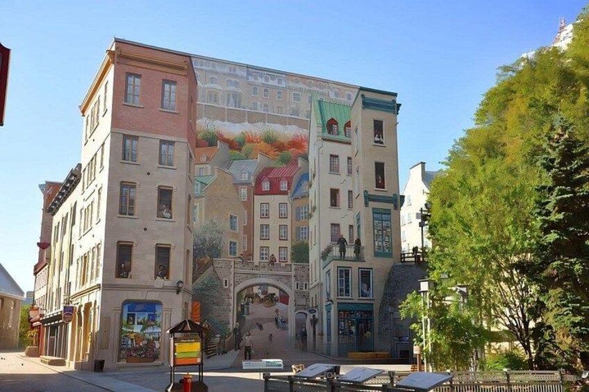 A multi-story building with a colorful mural on its facade, a street with benches, and a tree in the foreground. 3