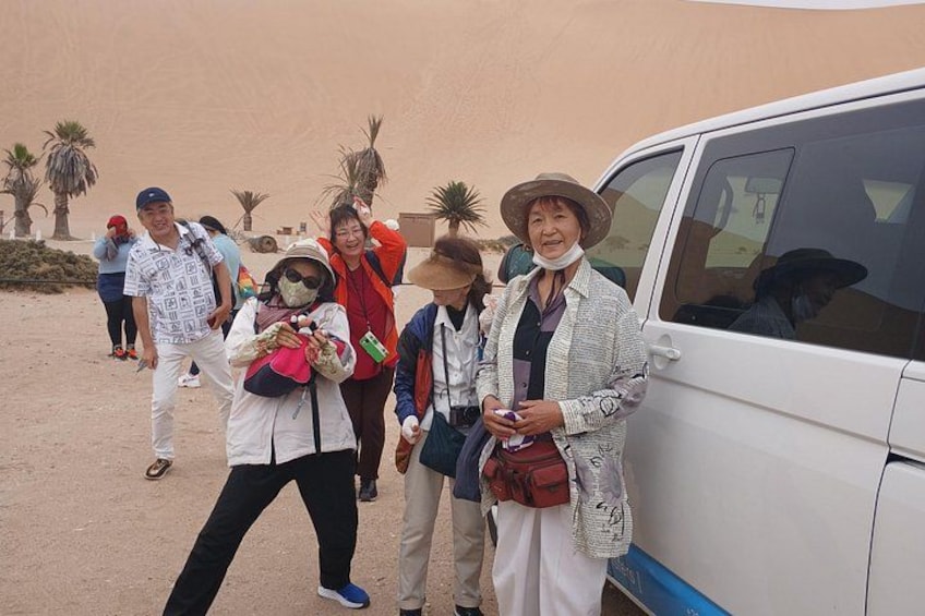 A group of people standing in a desert-like environment with palm trees in the background, a white vehicle parked to the right, and a sandy ground. 4