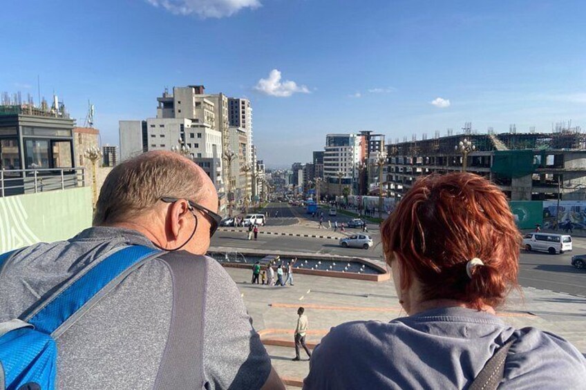 Twee personen kijken uit over een stadsgezicht met gebouwen en een drukke straat. 4