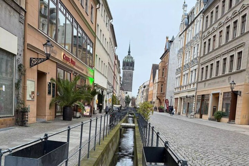 A cobblestone street with buildings on either side, a canal in the center, and a tower in the background. 1