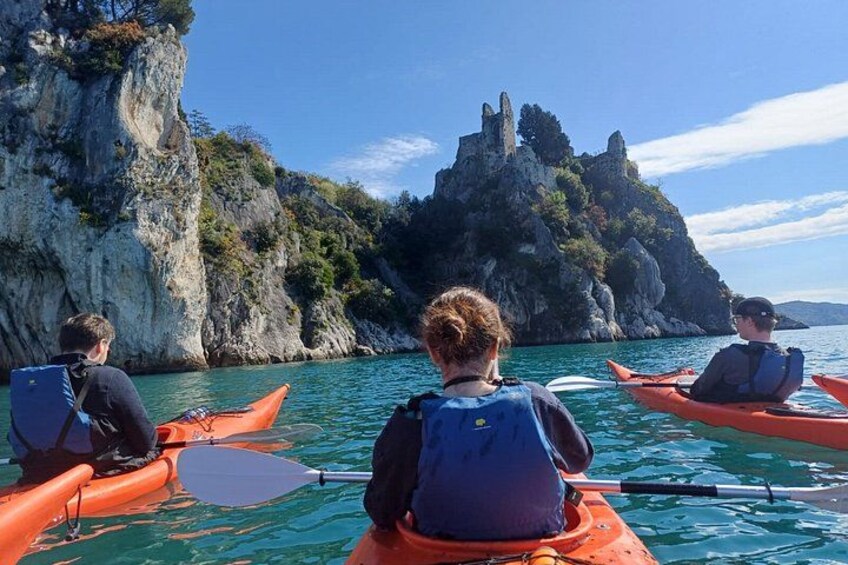 Three people kayaking on a calm body of water with rocky cliffs and a castle-like structure in the background. 1