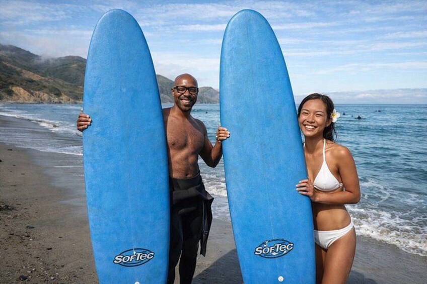 Two people holding blue surfboards on a beach with ocean waves in the background. 1