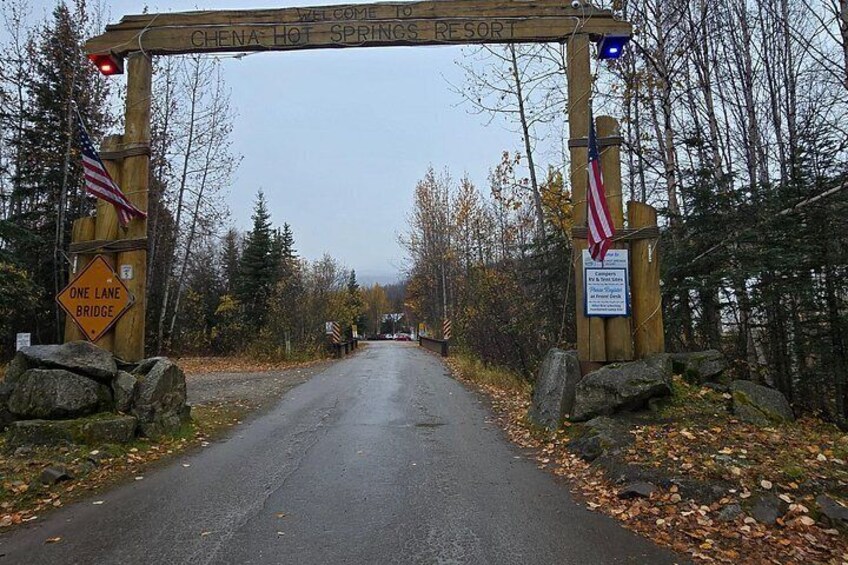 A wooden archway with 'Welcome to Chena Hot Springs Resort' written on it, flanked by American flags and a one-lane bridge sign. 4