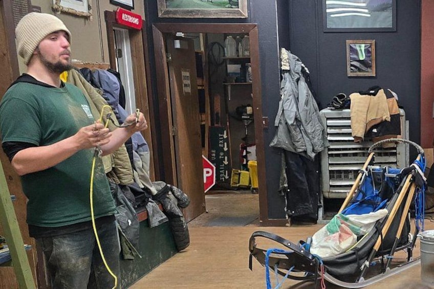 Un homme vêtu d’une chemise verte et d’un bonnet, se tient à côté d’une luge munie d’une attache. On aperçoit des vêtements suspendus à un portant et une porte sur laquelle est apposée une pancarte indiquant « POUSSER POUR SORTIR ». 2