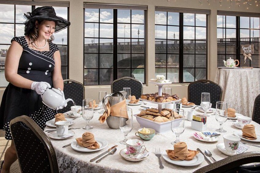 A woman pouring tea at a table set for a meal, with pastries and a sign reading 'Guest Book' in the background. 1