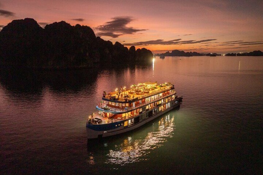 A large, illuminated river cruise ship with multiple decks and lights, sailing near rocky islands at dusk. 1