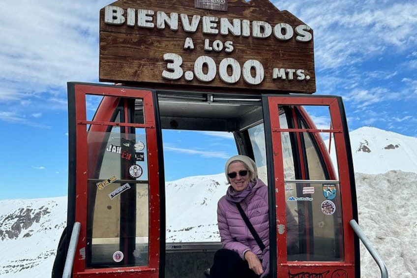 A wooden sign with 'BIENVENIDOS A LOS 3000 MTS.' text, a red and glass structure, and a snowy mountain background. 2