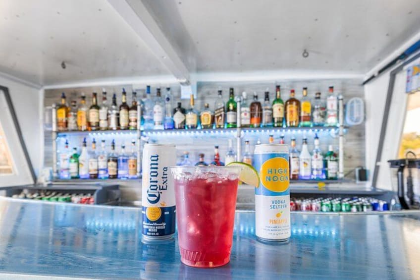 En bar med flasker af Corona Extra og HiNoon Vodka Seltzer, en rød drik med et limeskive, samt diverse flasker med alkohol i baggrunden. 2