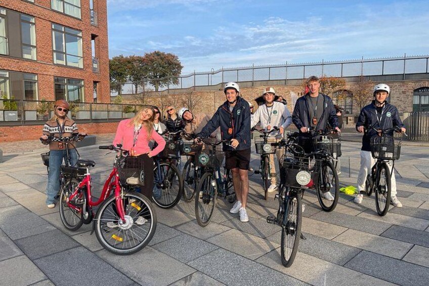 En gruppe mennesker med cykler i et udendørs miljø med en murstensbygning og en klar himmel. 3