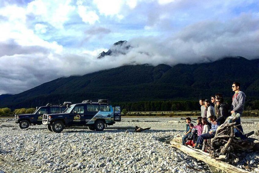 A group of people on a wooden raft near a truck on a gravel beach with mountains in the background. 4