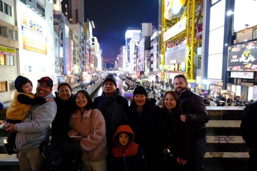 Un grupo de personas en una calle concurrida de noche, con edificios e indicaciones iluminadas al fondo. 2
