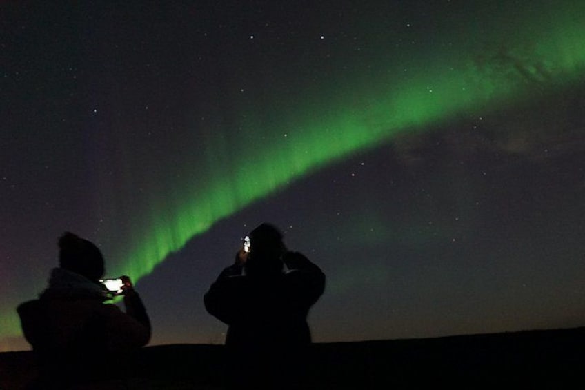 Two people silhouetted against a night sky, one holding a camera, and the Northern Lights in the background. 2