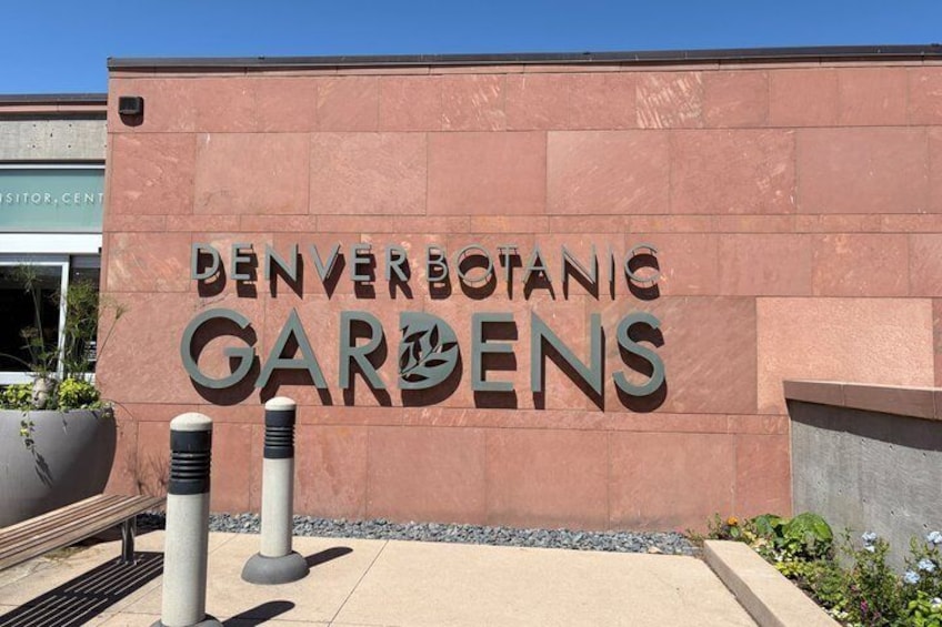 The Denver Botanical Gardens entrance with a large sign, a paved walkway, and a planter with plants. 2