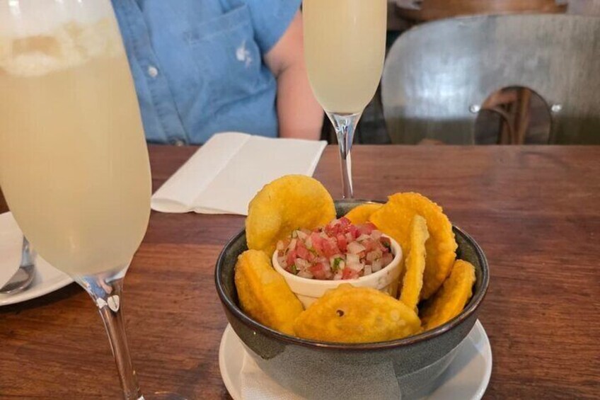 Un plato de plátanos fritos con una porción de salsa, acompañado por dos vasos de una bebida clara, probablemente un cóctel o un refresco, sobre una mesa de madera. 2